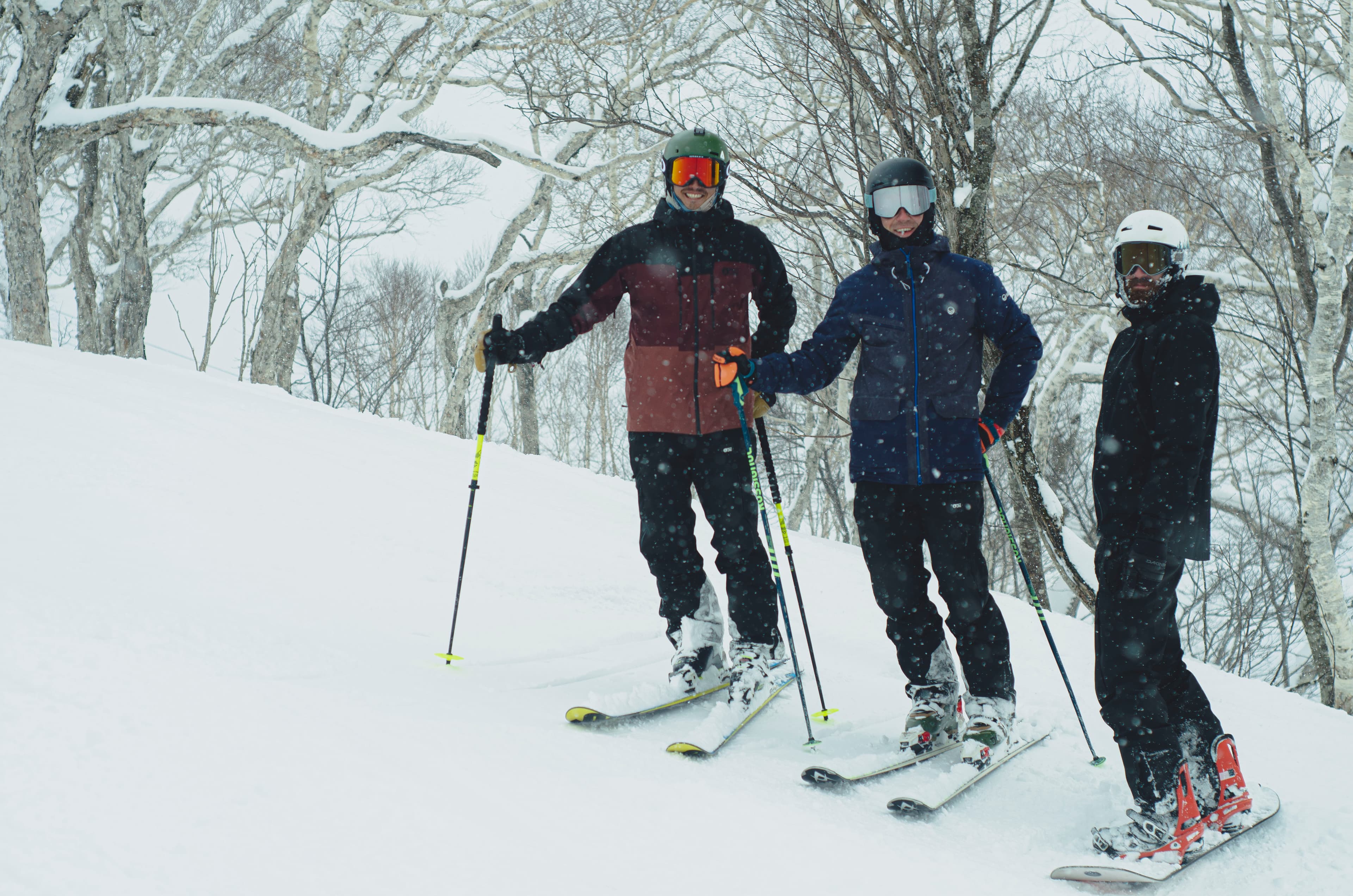Two skiers and a snowboarder pose on a snowy mountain slope during light snowfall.