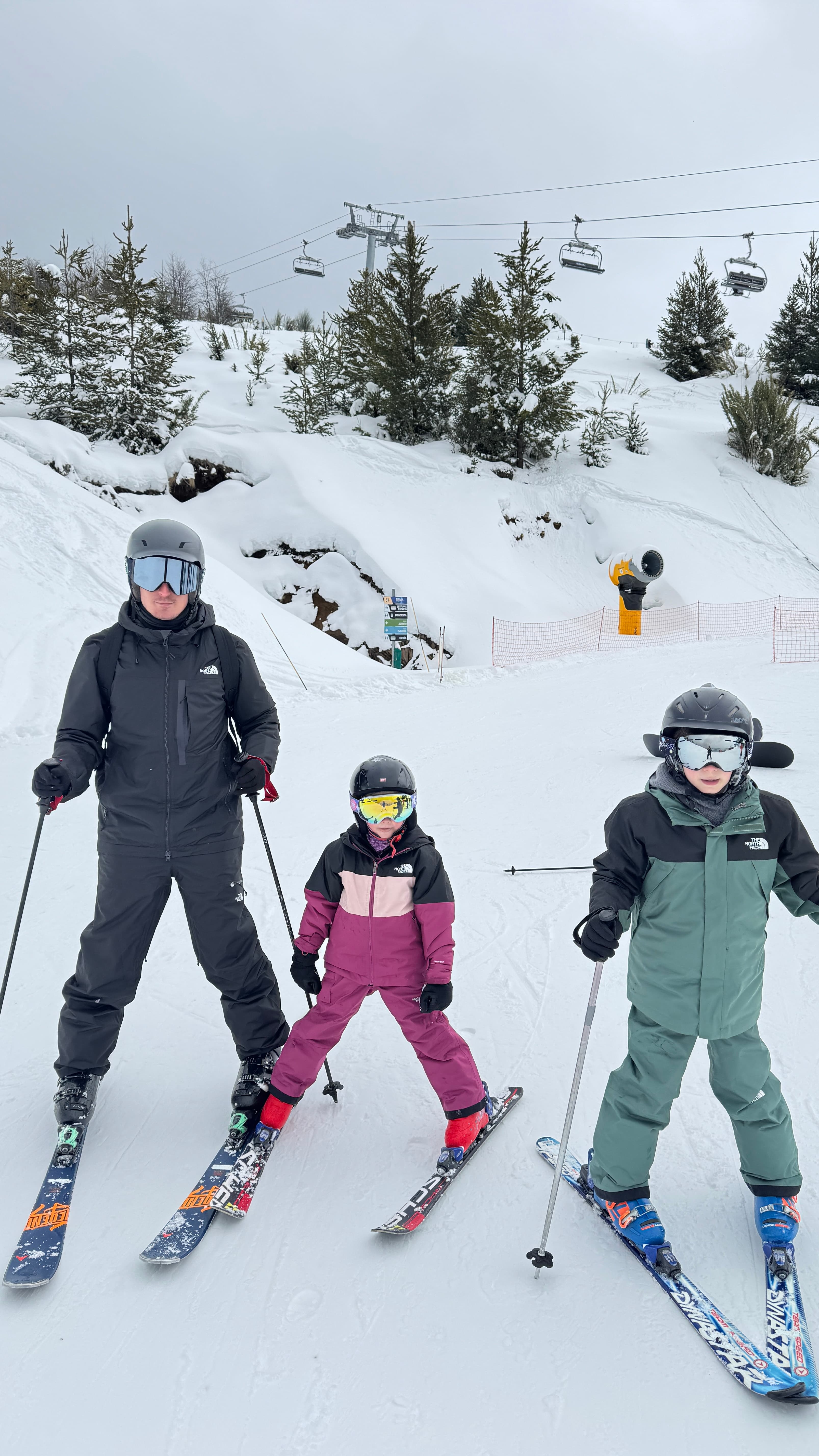 Man and two children in ski gear stand on a snowy mountain slope with chairlifts.
