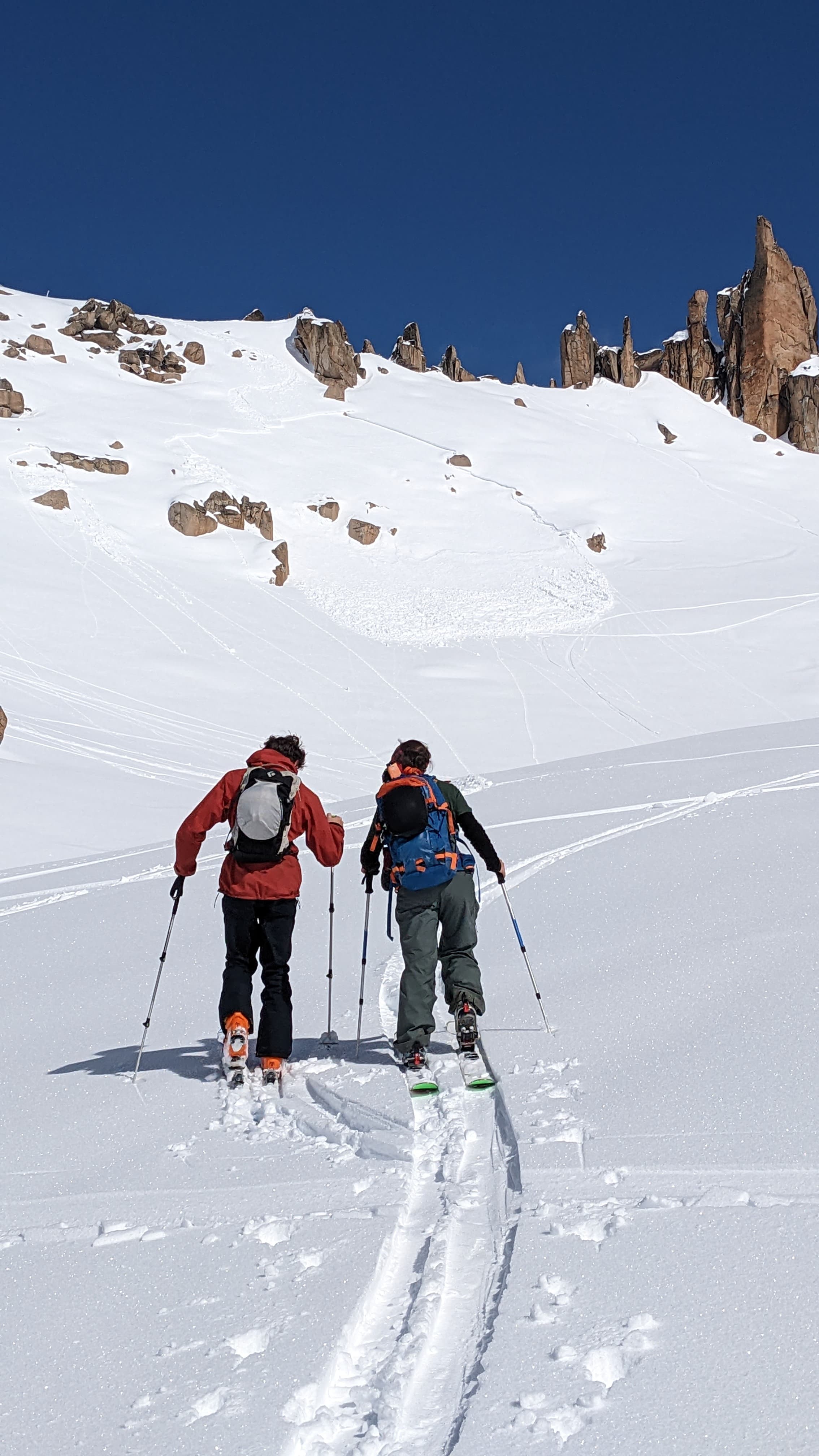 Two backcountry skiers trek up a snowy mountain slope toward rocky peaks under blue skies.