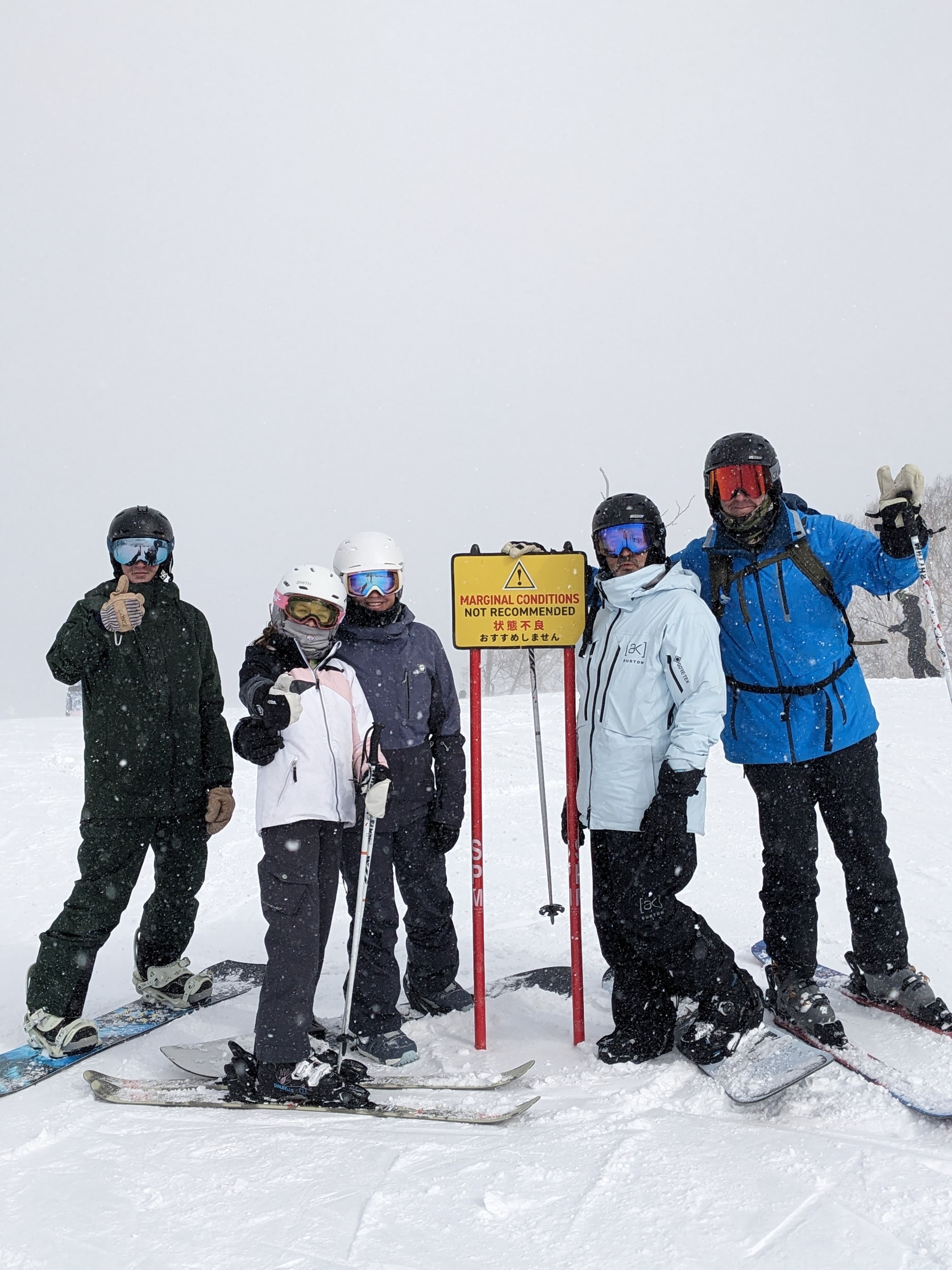 Five skiers and snowboarders pose next to a marginal conditions sign during a heavy snowfall.